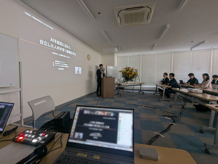 Presenter giving a talk at a podium about using AI in management, in a conference room with a projected slide behind and an audience seated at tables.