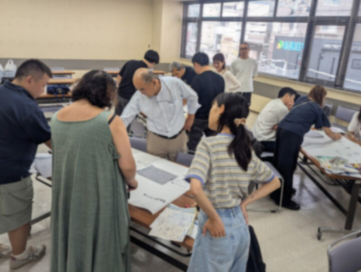 Group of adults gathered around tables in a classroom, looking over large sheets of paper and documents during a workshop.