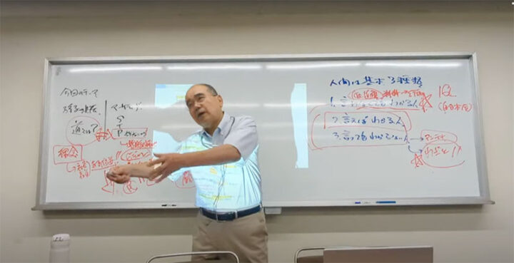 Man in a light blue shirt giving a lecture, gesturing toward a whiteboard filled with red handwritten notes in Chinese.