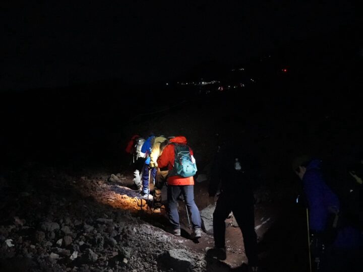Hikers with colorful backpacks walking a rocky mountain trail at night, illuminated by a flashlight/torchlight.