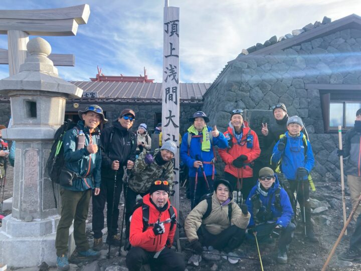 Group of hikers posing with thumbs up at a mountain summit marker, flanked by a stone lantern and torii in clear weather.