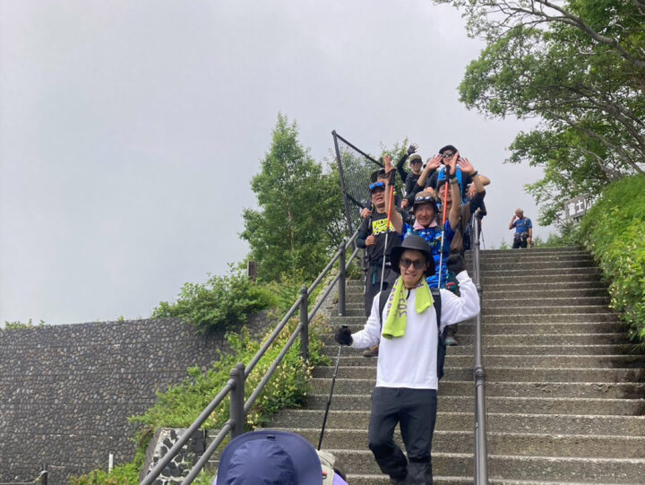 Group of hikers on a stone staircase, waving at the camera and wearing backpacks and hats during an outdoor trip.
