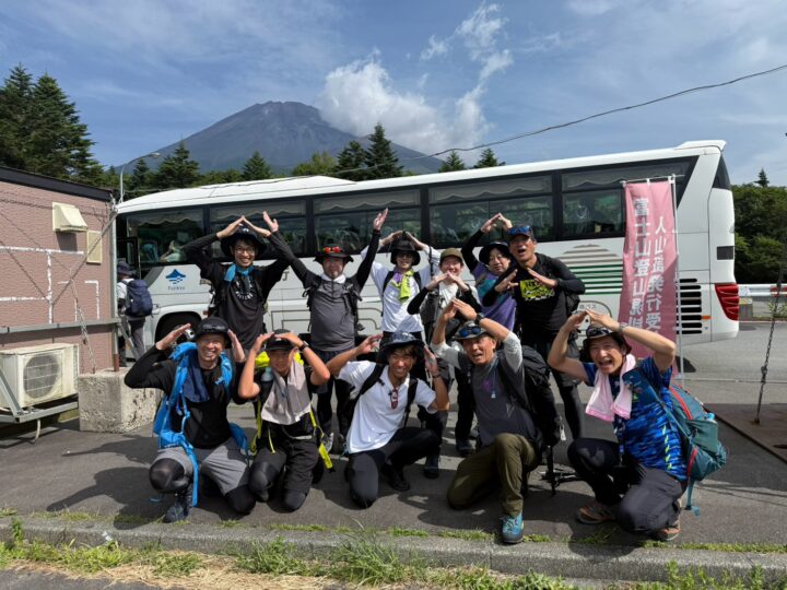 Group of hikers posing for a photo in front of a parked tour bus, with a mountain and trees in the background.