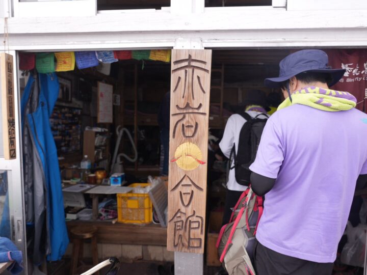 Vertical wooden sign with Japanese characters at a small shop entrance; customers browse inside, backpacks visible behind.