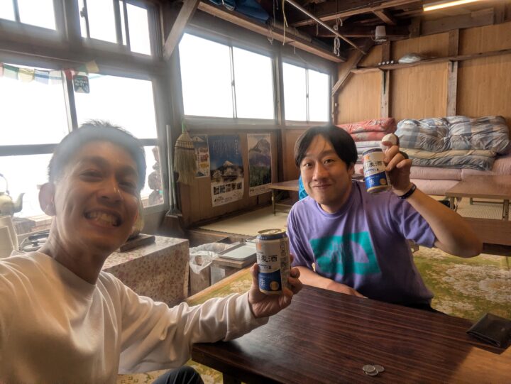 Two men in a rustic room clinking cans of beer at a wooden table, smiling at the camera with stacked cushions and windows in the background.