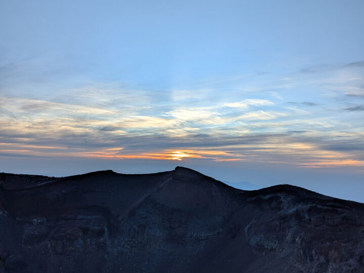 Sunrise over a dark, rugged volcanic landscape with a blue sky and orange horizon.